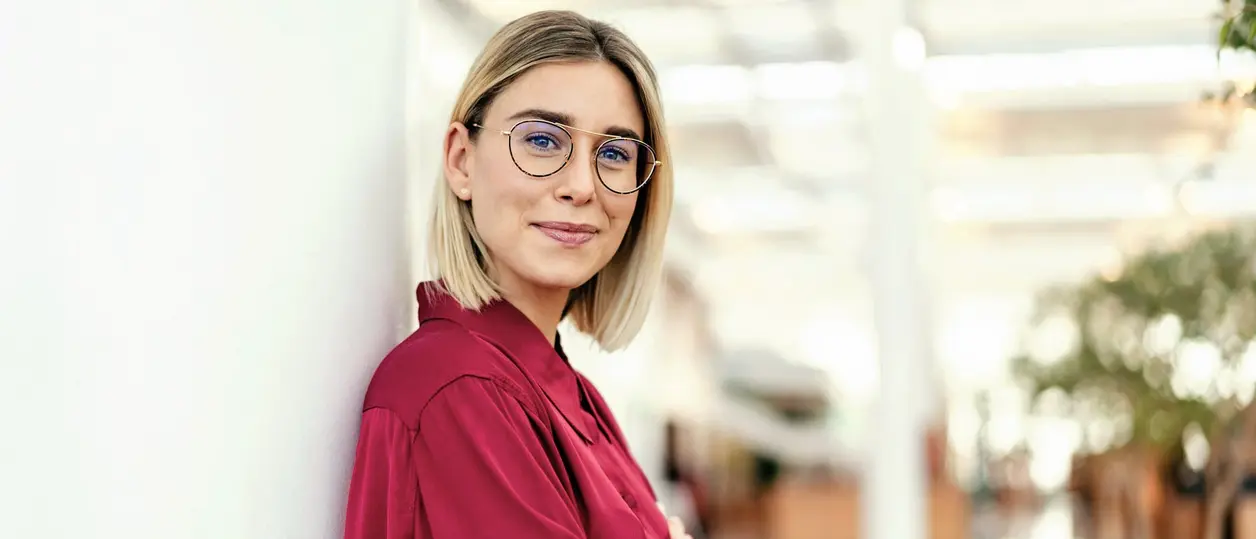 Junge Frau mit Brille lehnt entspannt an einer Wand im hellen Büro.