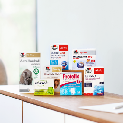 A collection of dietary supplements displayed on a wooden shelf. Products include "Anti-Hairball Complex" for cats, "Magnesium 400," "Skin-Hair-Nail," "Protefix Active Cleanser," and "Pure-3" Omega-3 capsules. The packaging is colorful, highlighting various health benefits.