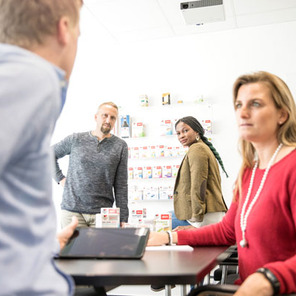 Four people are engaged in a discussion in a well-lit office. One person sits at a table with a tablet, while the others stand and lean in, looking toward the seated individual. A display of products is visible on the wall behind them.