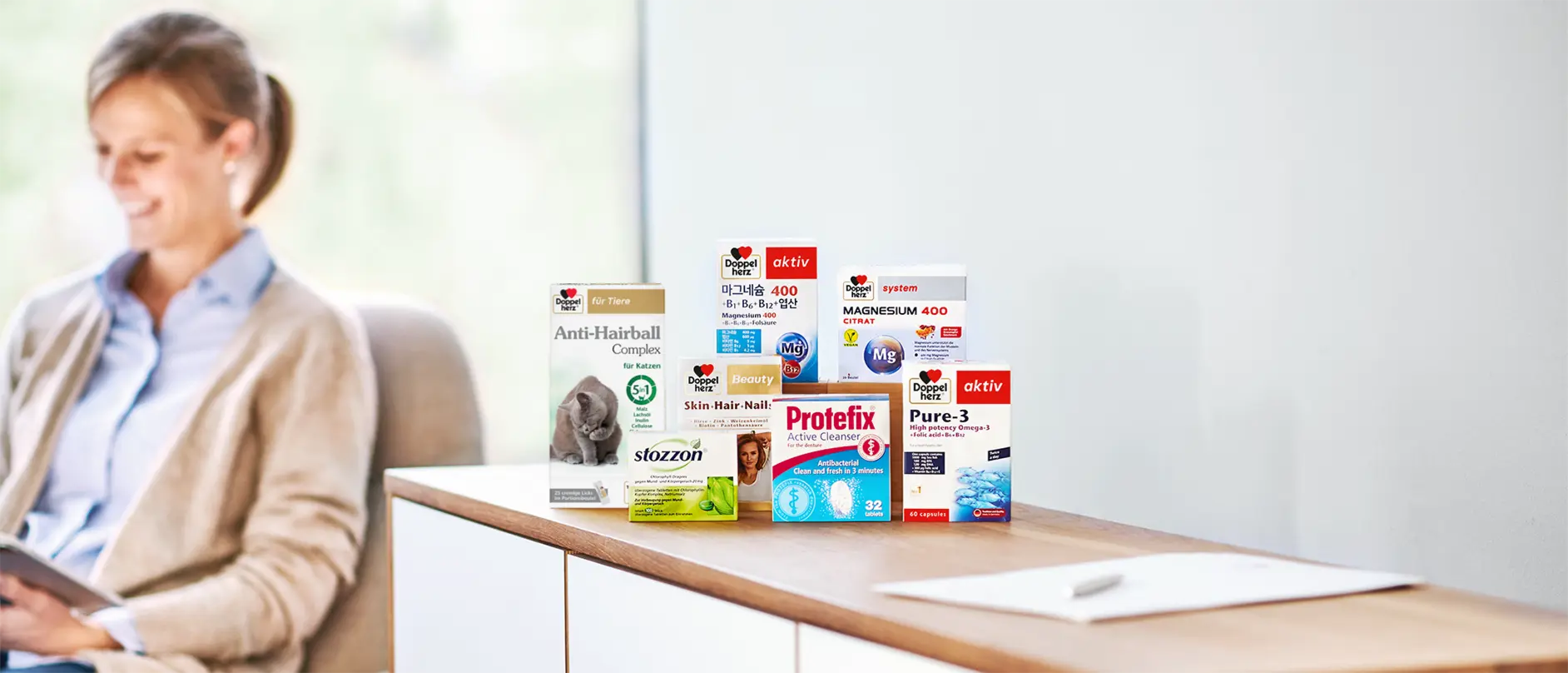 An assortment of health products displayed on a wooden surface. Items include an anti-hairball complex for cats, magnesium supplements, skin and hair support, an antibacterial cleaner, and omega-3 capsules. A woman is sitting in the background, smiling and reading on a tablet.