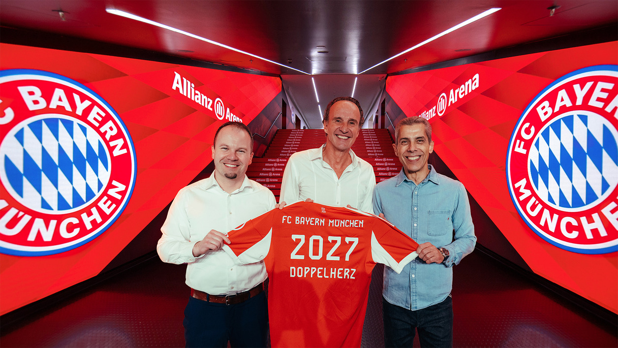 Three individuals are standing together, smiling and holding up a red FC Bayern München jersey displaying "2027" and "DOPPELHERZ." They are positioned in a well-lit corridor decorated with large team logos and branding for Allianz Arena, suggesting a celebratory event or announcement.